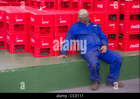 Ein Mann im blauen Overall schläft gelehnt einen Stapel von Coca Cola Kisten Stockfoto