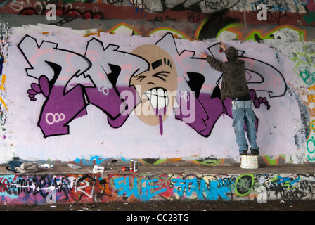 Tagging - Kapuzen-junger Mann Spray Malerei Graffiti unter Brücke. Stockfoto