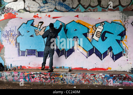 Tagging - Kapuzen-junger Mann Spray Malerei Graffiti unter Brücke. Stockfoto