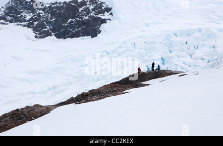Touristen sitzen auf einem Felsvorsprung Look neben einem Gletscher über Neko Harbour, Antarktis Stockfoto