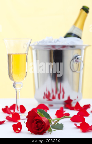 Foto von einer roten Rose und einem Glas Champagner auf einer weißen Leinen Tischdecke mit einer Flasche Champagner in einem Eiskübel Stockfoto