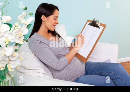 Foto von einer schwangeren Frau zu Hause sitzt in einem Sessel, schreiben eine Liste der möglichen jungen und Mädchen Namen für ihr Baby. Stockfoto