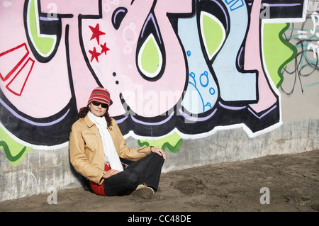 Frau mit einem Hut und Schal sitzen gegen eine Wand gekreuzt-legged bedeckt in graffiti Stockfoto