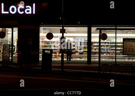 Mann in einem kleinen Supermarkt einkaufen, nach Einbruch der Dunkelheit, fotografiert von außerhalb Stockfoto