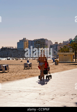 junge Mutter in Bikini gekleidet schiebt Kinderwagen entlang der Promenade am Playa Levante, Benidorm, Costa Blanca, Spanien Stockfoto