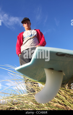 Eine Surfer mit Händen in den Taschen steht über seinem Surfbrett vor blauem Himmel. Stockfoto