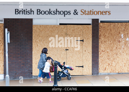 Frau mit einem Kind schiebt einen Kinderwagen vorbei an einem vernagelten Shop. Stockfoto
