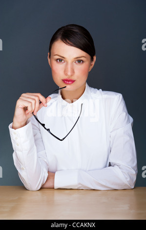 Schöne europäische junge solide Geschäftsfrau mit Brille vor grauem Hintergrund sitzen Stockfoto