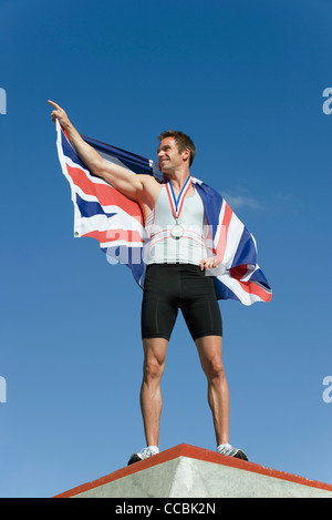 Männlicher Athlet auf Siegertreppchen, britische Fahne hochhalten Stockfoto