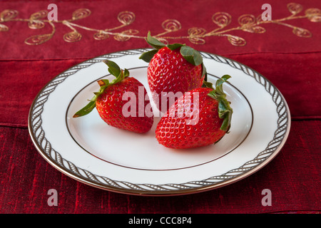 Drei rote Erdbeeren mit grünen Blättern auf einem weißen feinen Porzellanteller mit Silberschmuck auf der Felge. Roten strukturierten Hintergrund. Stockfoto