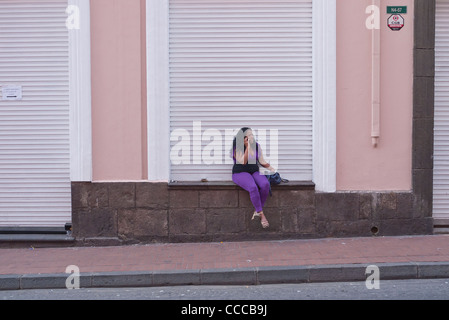 Eine 40 Jahre alte weibliche Straße Walker sitzt auf einer Mauer vor einem geschlossenen laden während des Gesprächs auf ihrem Handy in Quito, Ecuador. Stockfoto