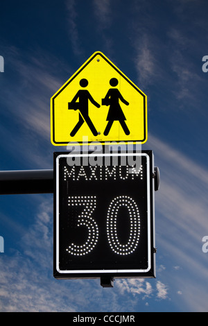 Ein elektrischer 30km/h Höchstgeschwindigkeit Zeichen, unter einem Zeichen, die den Beginn einer Schule Zone gegen einen blauen Himmel & weiße Wolken zeigen Stockfoto