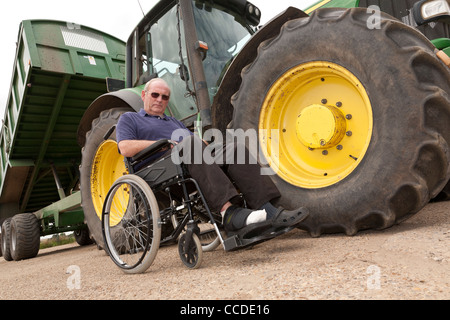 Ein Mann in einem Rollstuhl neben einer landwirtschaftlichen Zugmaschine und Anhänger Stockfoto