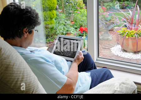 Ältere ältere Rentnerin Frau hält Apple iPad digitalen Tablet-Computer Zuhause allein am Fenster sitzen im Sessel Sommergarten draußen England, Großbritannien Stockfoto