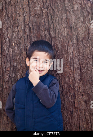 Kleiner Junge auf der Suche nach links vor einem Baum mit Rinde Textur im Hintergrund beißen auf seinen Finger mit einem schüchternen Ausdruck Stockfoto