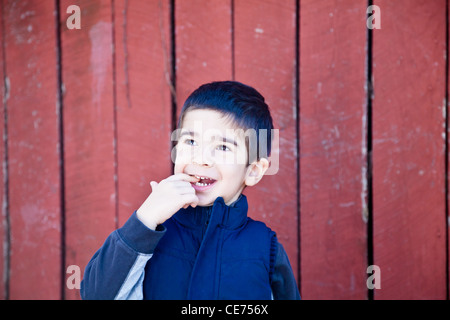 Kleiner Junge auf der Suche nach oben und nach links vor roten strukturierten Hintergrund beißen auf seinen Finger mit einem Staunen Ausdruck. Stockfoto