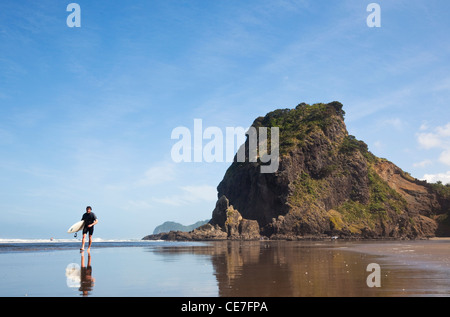 Surfer am Strand mit Lion Beach im Hintergrund. Piha, Waitakere Ranges Regional Park, Auckland, Nordinsel, Neuseeland Stockfoto