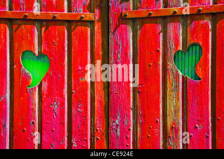 Zwei Herzformen geschnitten in einem roten Holztor enthüllt Grünholz hinter, symbolisch für Neid, Eifersucht, Leidenschaft Stockfoto