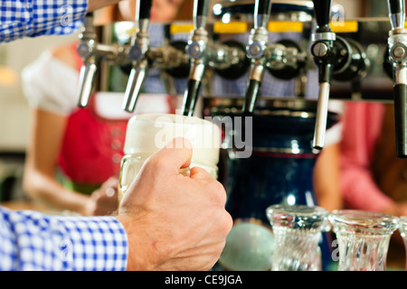 Mann ein Bier vom Hahn auf einem Kegerator im Pub oder Inn, Menschen in bayerischer Tracht Zeichnung stehen im Hintergrund Stockfoto