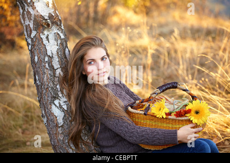 Schöne junge Frau mit einem Korb voller Blumen sitzen im Schatten eines Baumes im Herbst. Stockfoto