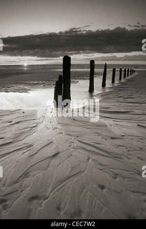 Die Sonne versinkt hinter einer Reihe von alten hölzernen Buhnen an einem Sandstrand in Brancaster, Norfolk Stockfoto