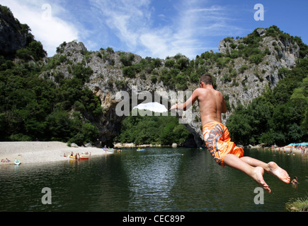 Ein Junge springt ins Wasser der Ardèche. Im Hintergrund die Brücke von Vallon Pont d ' Arc in Südfrankreich. Stockfoto