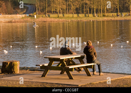 Paar (Mann & Frau) Sitzen entspannen, auf Picknick Tisch Sitzbank mit Blick auf Wasser an einem sonnigen Tag - malerische Swinsty Reservoir, Yorkshire, England, UK. Stockfoto