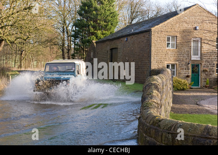 Überschwemmung - Land Rover Defender Fahren & Spritzwasser durch tiefe Hochwasser auf überfluteten Landstraße nach schweren Regenfällen - North Yorkshire, England, Großbritannien Stockfoto