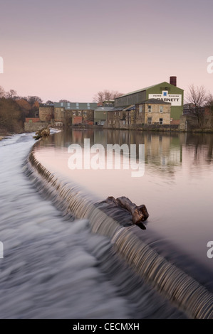 Fließendes Wasser von River Wharfe Kaskadierung über Wehr unter rosa Sonnenuntergang Himmel, historische Granat der Papierfabrik jenseits - Otley, West Yorkshire, England, UK. Stockfoto