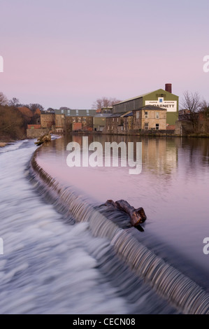 Fließendes Wasser von River Wharfe Kaskadierung über Wehr unter rosa Sonnenuntergang Himmel, historische Granat der Papierfabrik jenseits - Otley, West Yorkshire, England, UK. Stockfoto