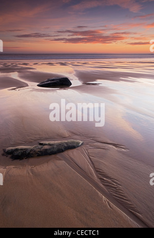 Dawn wirft rosa Licht und Reflexionen über Felsen links von der Flut an einem nassen Sandstrand. Stockfoto