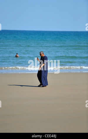 Ein einsamer omanischer Mann seine Telefonnummer überprüfen während des Gehens auf dem breiten Sandstrand am Yiti; Muscat, Oman Stockfoto