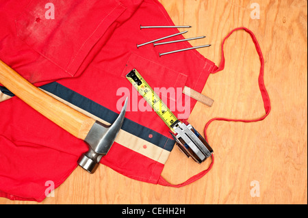 Werkzeuge für die Holzbearbeitung wie einen Hammer, Maßband, Bleistift Tischler und alte rote Schürze auf ein Stück Sperrholz Planke Stockfoto