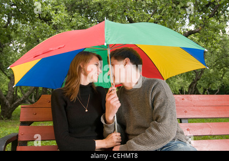 Glückliches junges Paar auf einer Bank. Unter einem bunten Regenschirm Stockfoto