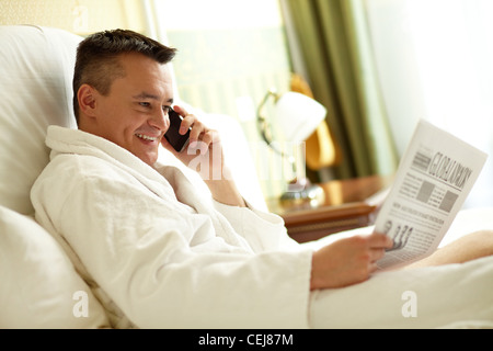 Gut aussehender Mann im Bademantel durch das Telefon sprechen, während Sie Zeitung im Bett lesen Stockfoto