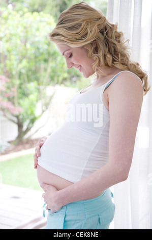 Schwangeren weiblichen gewellte blonde Haare, trägt eine weiße Weste und Hose, Stand am Fenster und Holding Beule, Lächeln, Stockfoto