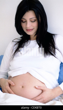 Hochschwangere Frauen lange brünette Haare tragen weiße Langarm t Shirt sitzen auf Kissen, stützte die Hände berühren Stockfoto