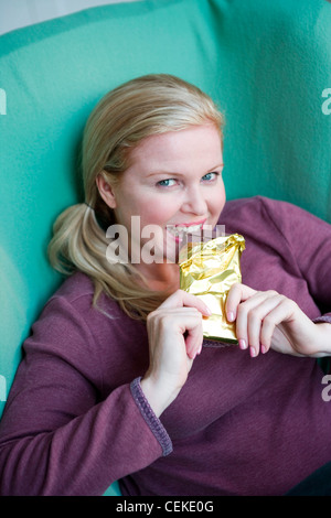 Nahaufnahme eines größeren weibliche blonde Haare aus Gesicht tragen lila Top sitzen im Sessel Haltestange Schokolade in beiden Händen Stockfoto