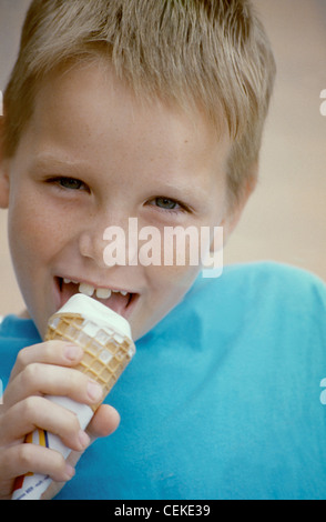 MClose, männliches Kind blonde Haare tragen blaue t-Shirt Essen Vanille Eiscreme-Kegel auf Kamera Lächeln zeigt Zähne Stockfoto