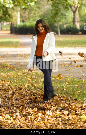 Weibchen tragen orange Rollkragen Pullover Creme Jacke und dunkelblaue Jeans spazieren im Park treten lässt in Luft Stockfoto