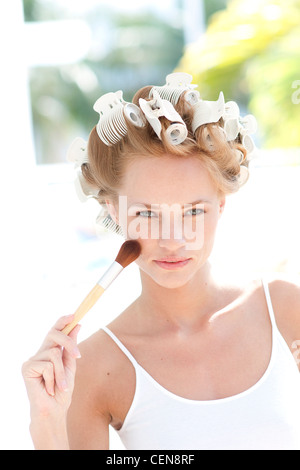 Weibliche rote Haare gewickelt auf weißen Lockenwickler, tragen eine weiße Weste, Make up Pinsel an ihre Wange halten ernsten, auf der Suche Stockfoto