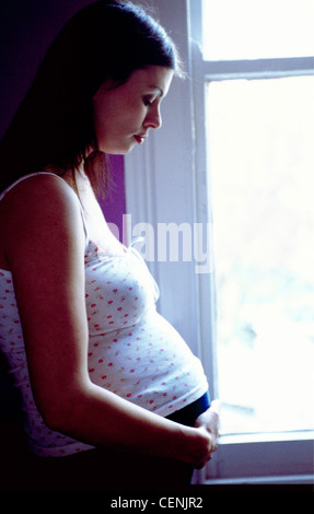 Profil von hochschwangere Frauen lange brünette Haare tragen florale Weste Top und Jeans im Fenster Hände unter Magen stehen Stockfoto