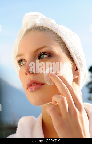 Weibchen tragen einen weißen Frottee Bademantel mit ihrem Haar in einem weißen Handtuch-Turban reiben Sahne in ihre Wange Stockfoto