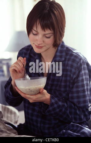 Weibchen mit kurze brünette Haare tragen blaue Tartan Pyjama sitzt auf Bett blickte Schneidersitz essen Müsli Stockfoto