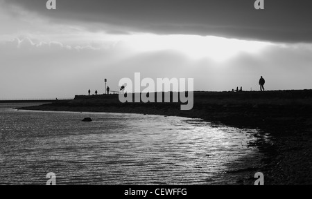 Ein Schuß von Menschen zu Fuß entlang der Küste der Bucht von Galway auf die Sonne durch die Wolken erzeugen Silhouetten zu brechen. Stockfoto