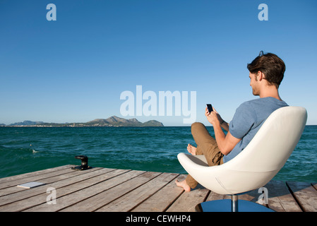 Junger Mann sitzt im Sessel See mit Handy, Seitenansicht Stockfoto