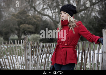 Weiblich, lange blonde Haare, trägt einen kurzen roten Zweireiher Mantel, schwarzen Hut, schwarze Handschuhe und Jeans, durch den Zaun stehen, Stockfoto