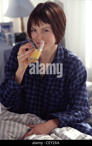 Weibliche kurze brünette Haare tragen blaue Tartan Pyjama sitzen im Bett trinken Orangensaft mit Glas in den Mund auf Stockfoto