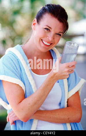 Frau nass brünette Haare vom Gesicht mit weißer Weste unter Shortsleeved Frottier Bademantel, Glas Wasser, Arm gefaltet Stockfoto