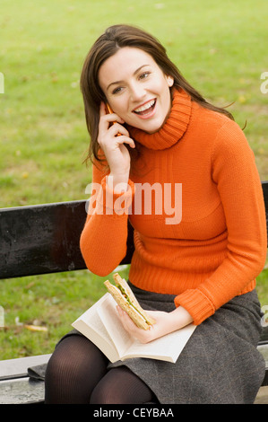 Weibchen tragen orange Rollkragen Pullover und grau Rock sitzt auf Park Bench Lesebuch und Sandwich zu essen und sprechen auf Stockfoto